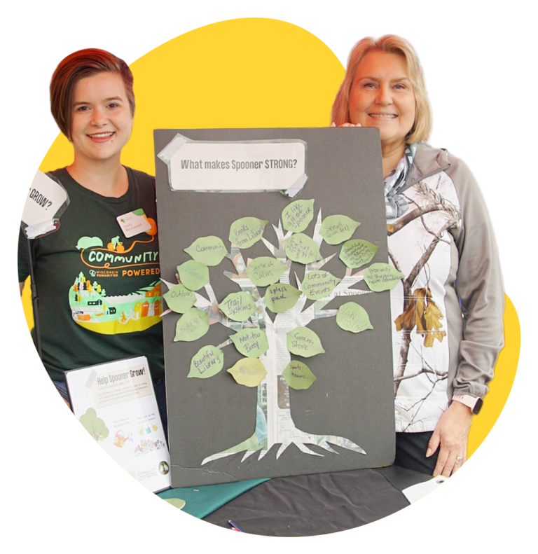 Two women stand in behind a board with a tree and the title "What makes Spooner STRONG?